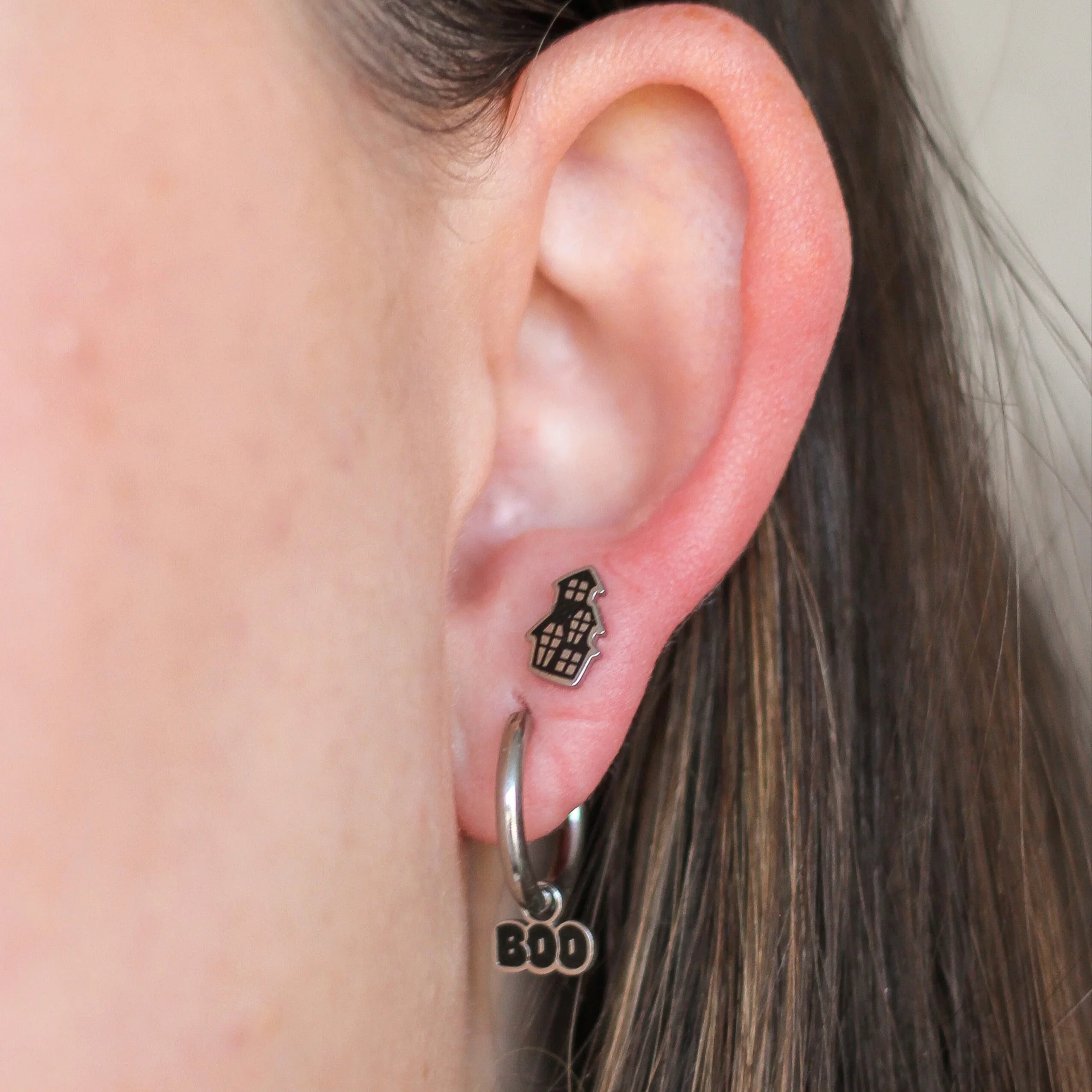 Close-up of an ear with hoop earrings and a small stud that is shaped like a black and silver haunted house.