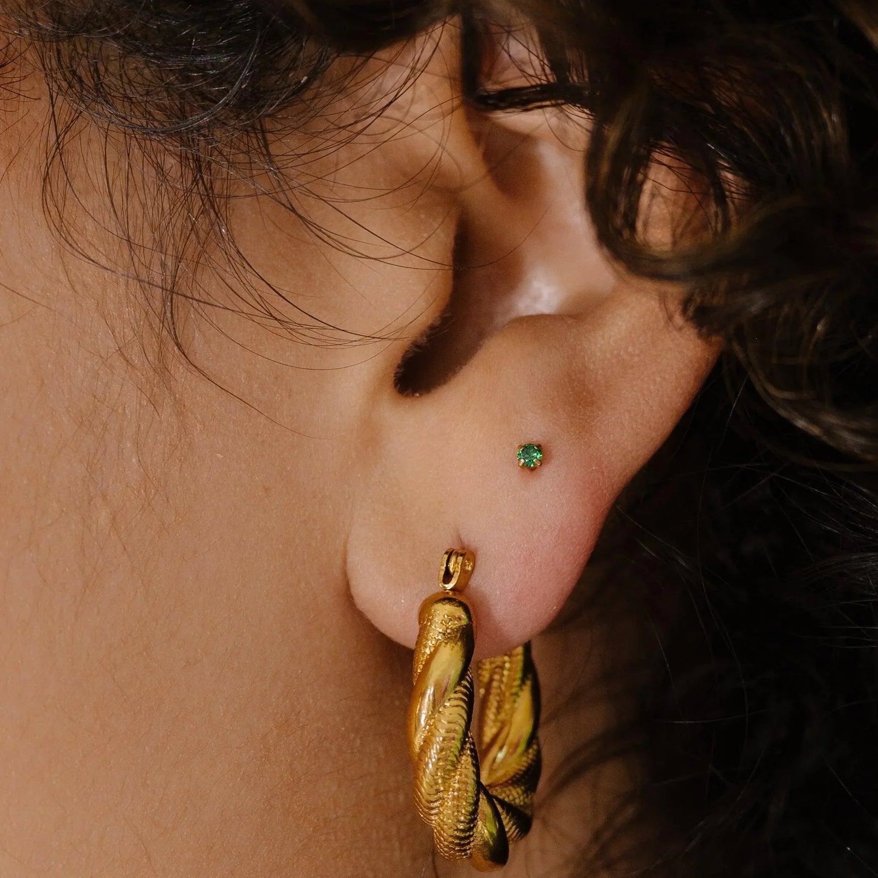 Close-up of an ear wearing a gold hoop earring and a tiny green CZ gemstone in the second lobe piercing.