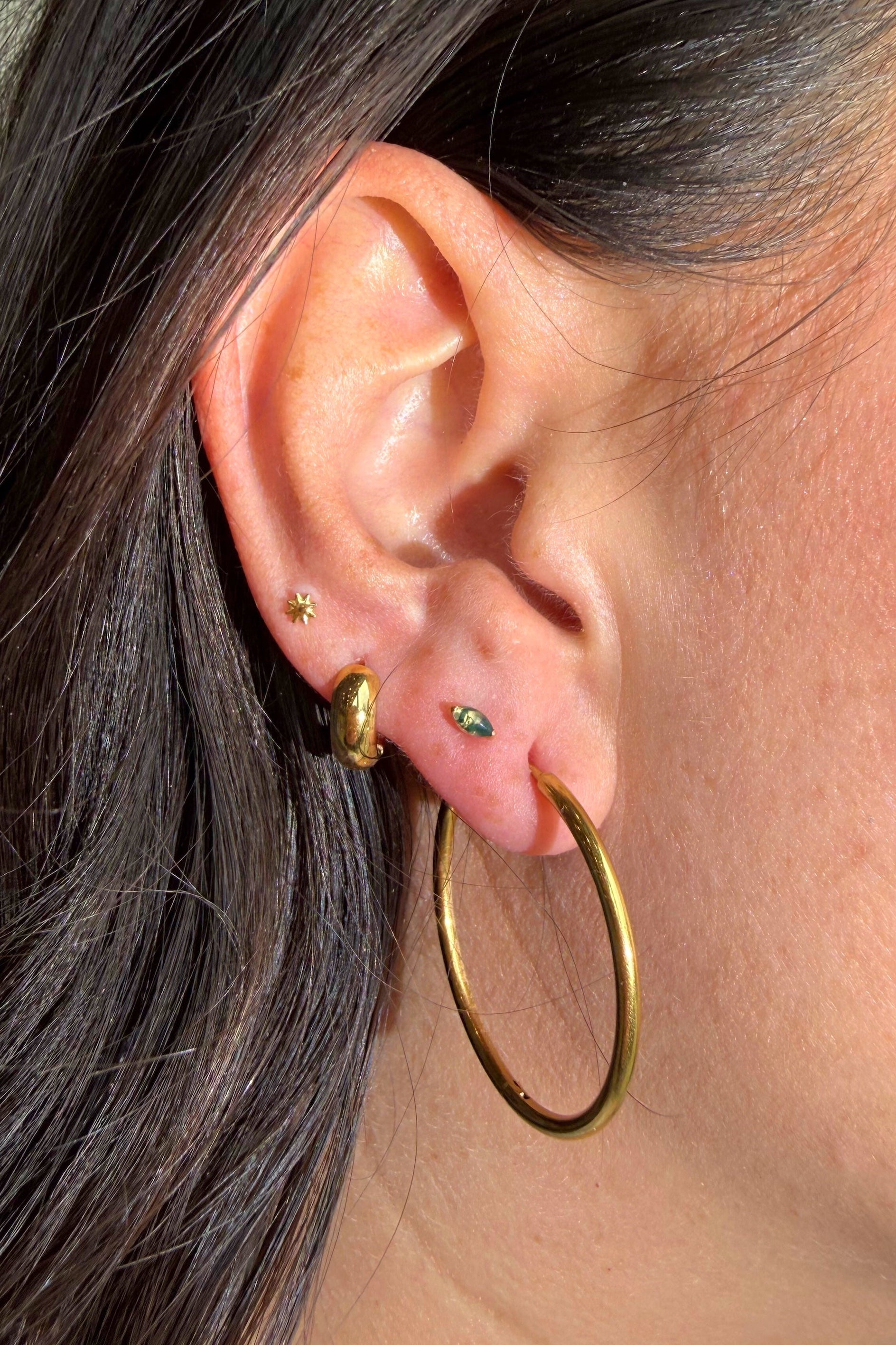 Close-up of an ear with gold hoop earrings and a small green earring.