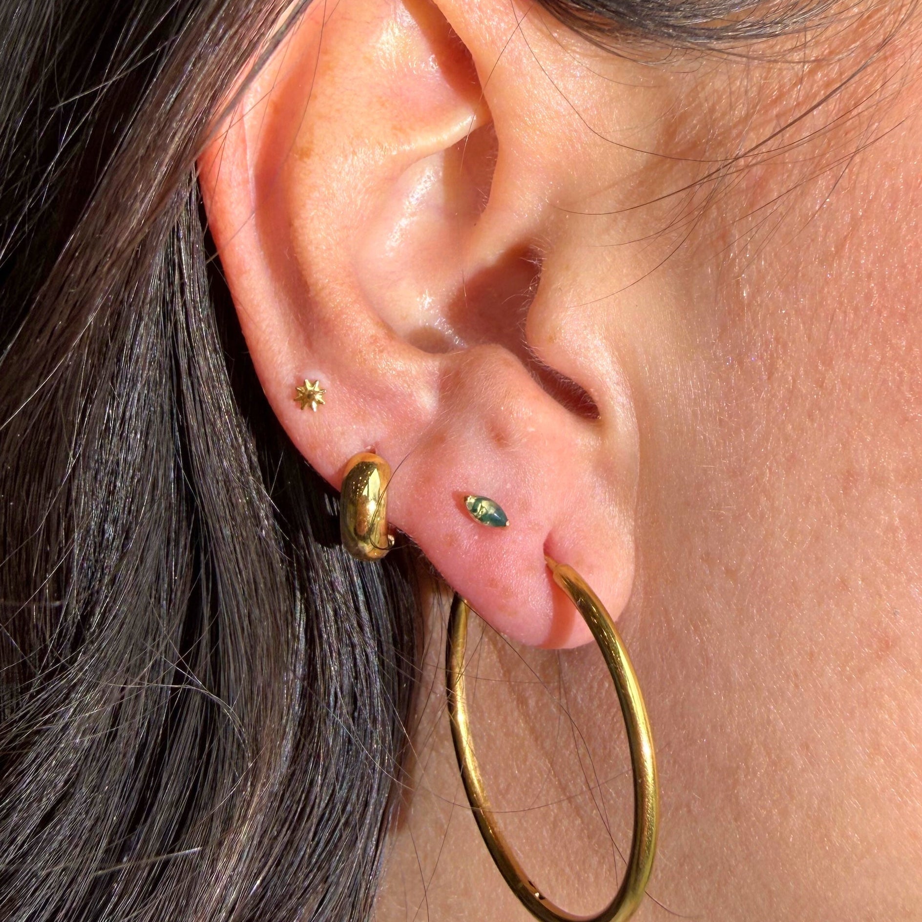 Close-up of an ear with gold hoop earrings and a small green earring.