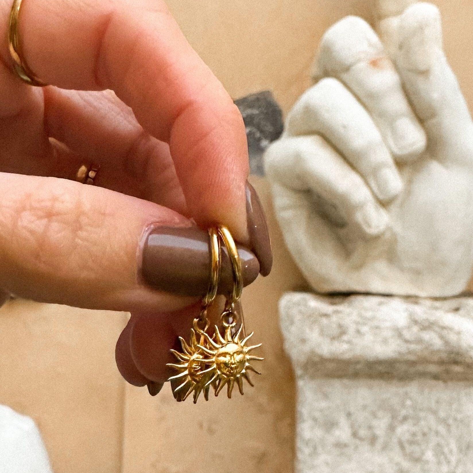 Gold earrings with a textured sun design held by a hand in front of a marble statue.