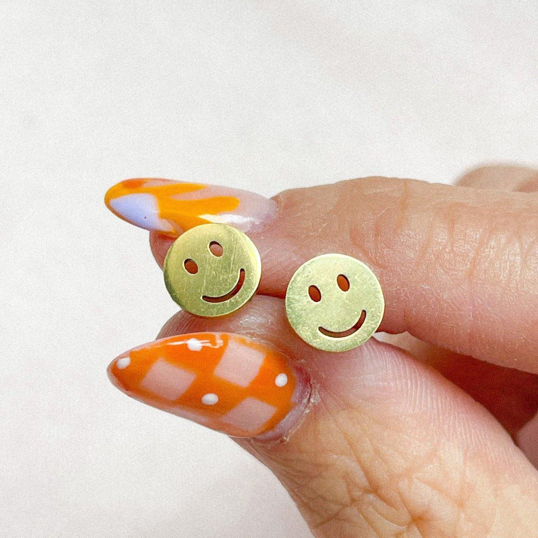 Gold smiley face earrings held between fingers with orange and white nail polish on a light background
