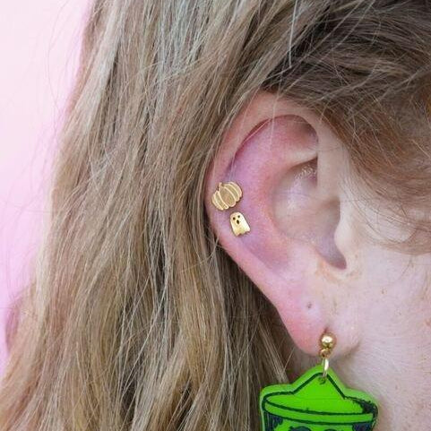 Close-up of an ear wearing a green pumpkin earring with a smiling face, against a pink background.